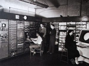 A black and white photo of a computer room with three men and a woman working on early computers